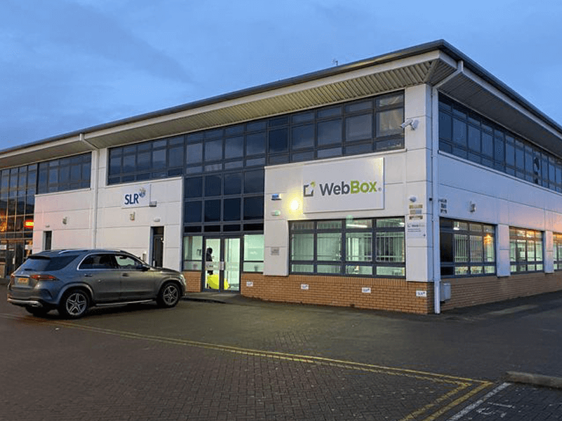 An office building with logos for SLR and WebBox on the facade. A parked car is visible in front of the building, which is illuminated by the evening light. Our new WebBox HQ makes the news.