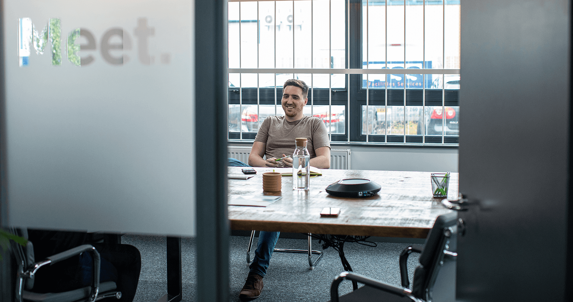 A person is sitting alone at a large table in a conference room with a glass door labeled "Meet." The room has large windows with bars, and there are office supplies on the table.