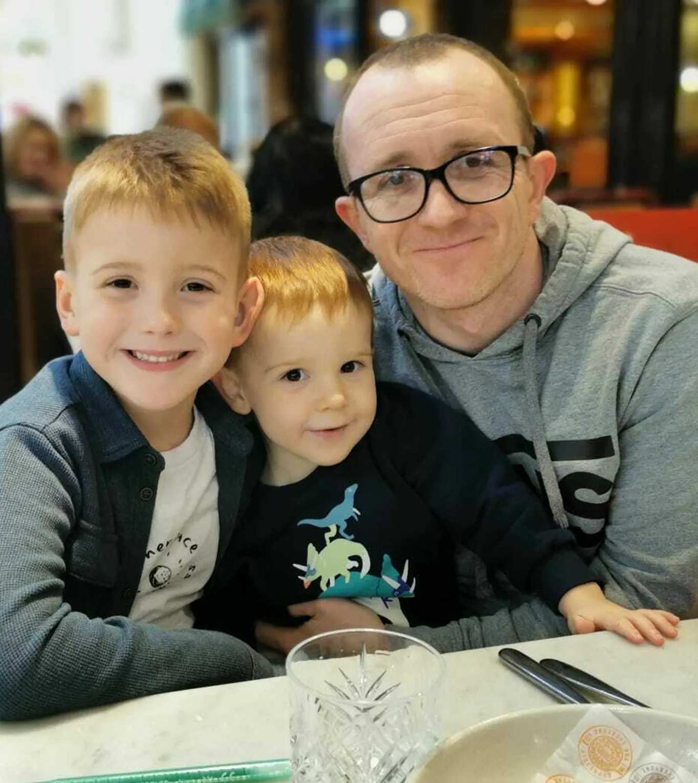 A man wearing glasses and a gray hoodie sits at a table with two young boys, all smiling.