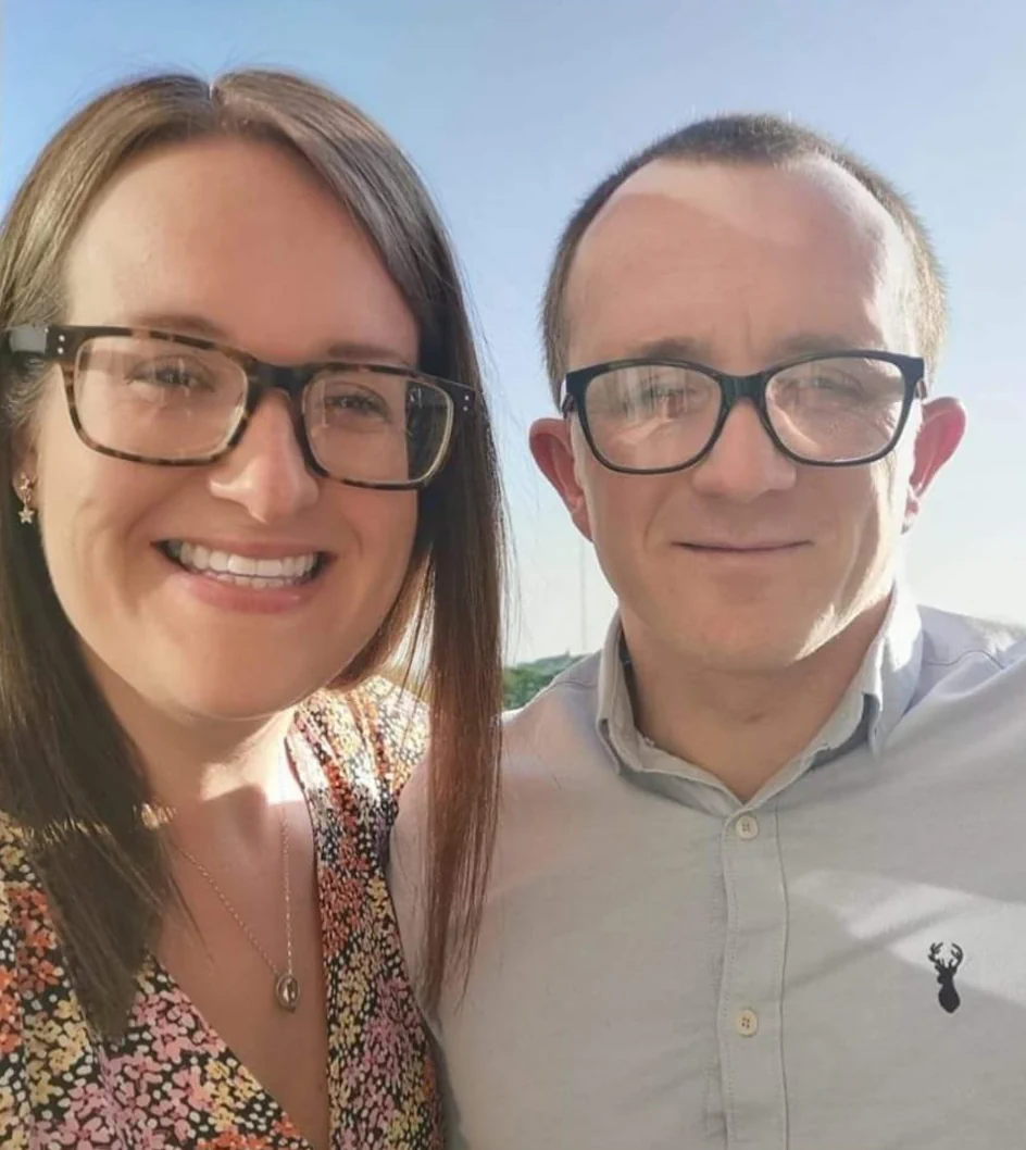 A woman and a man, both wearing glasses, smile at the camera. The woman is in a floral dress, and the man is in a light gray shirt with a dark emblem. They are outdoors with a clear sky in the background.