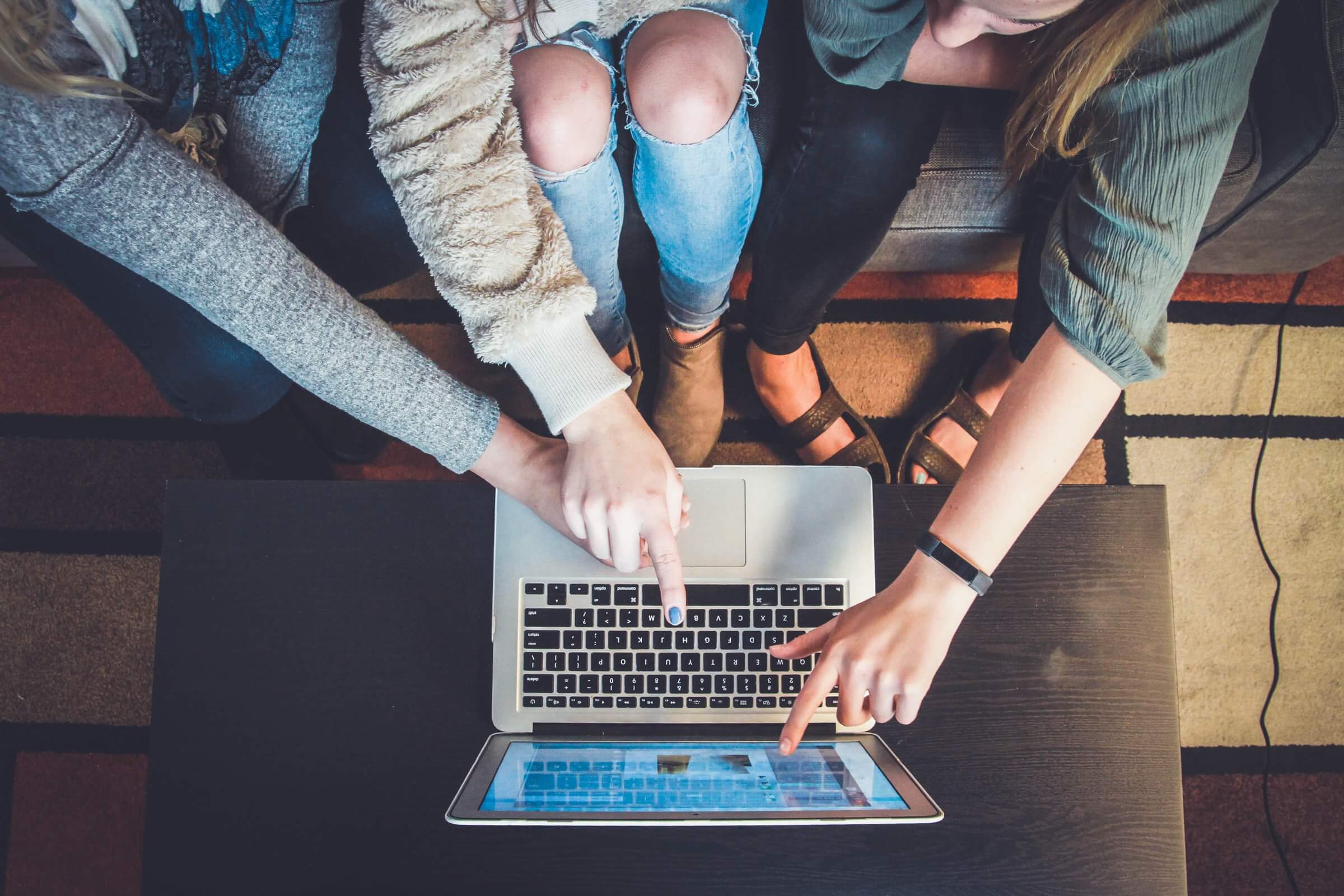 Three people sit at a table using a laptop. They are pointing at the screen and keyboard, suggesting collaboration or sharing information.