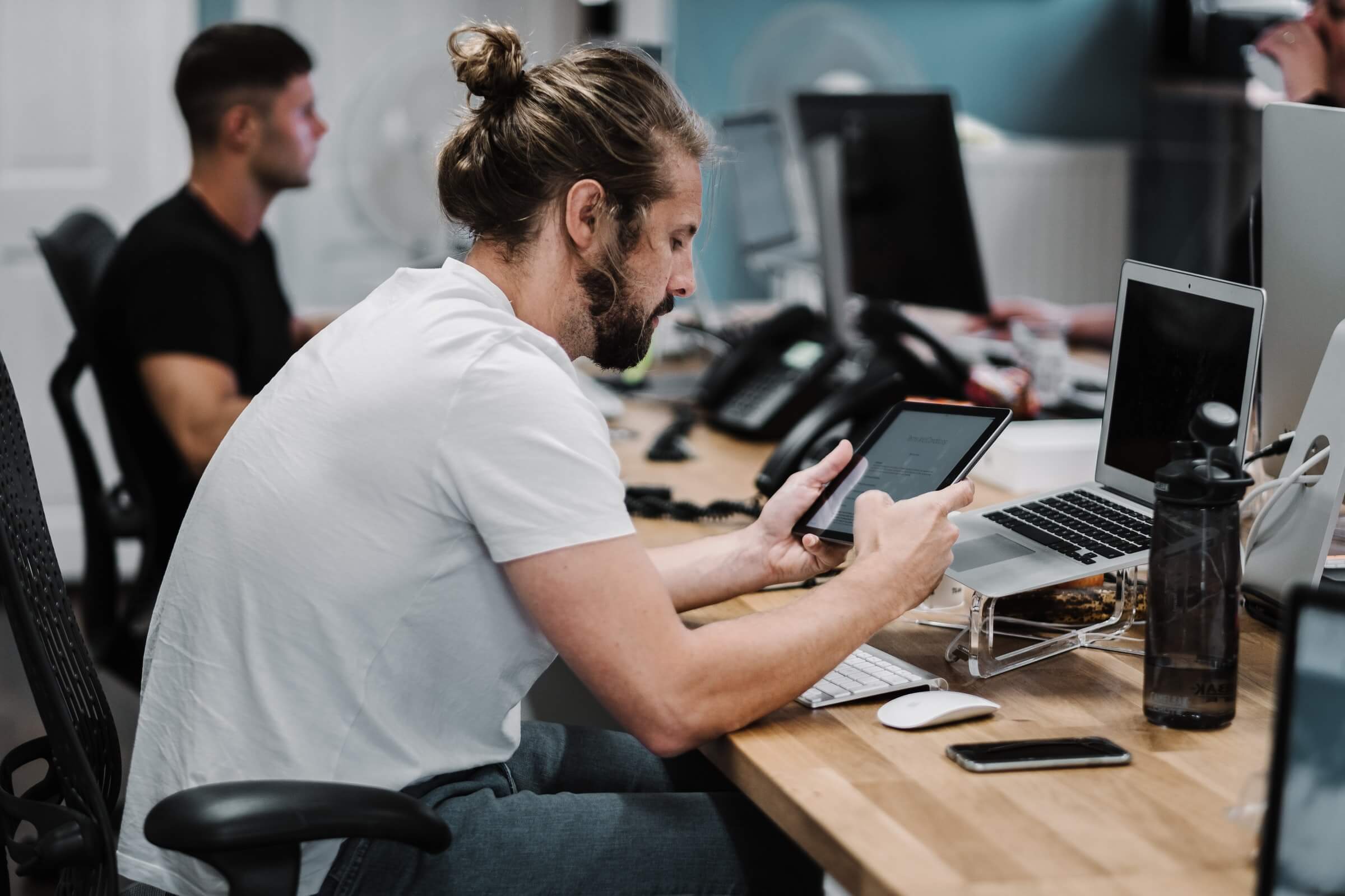 Man with a beard and a bun seated at a desk using a tablet, surrounded by a laptop, smartphone, and water bottle in an office environment. Another person works at a desk in the background.