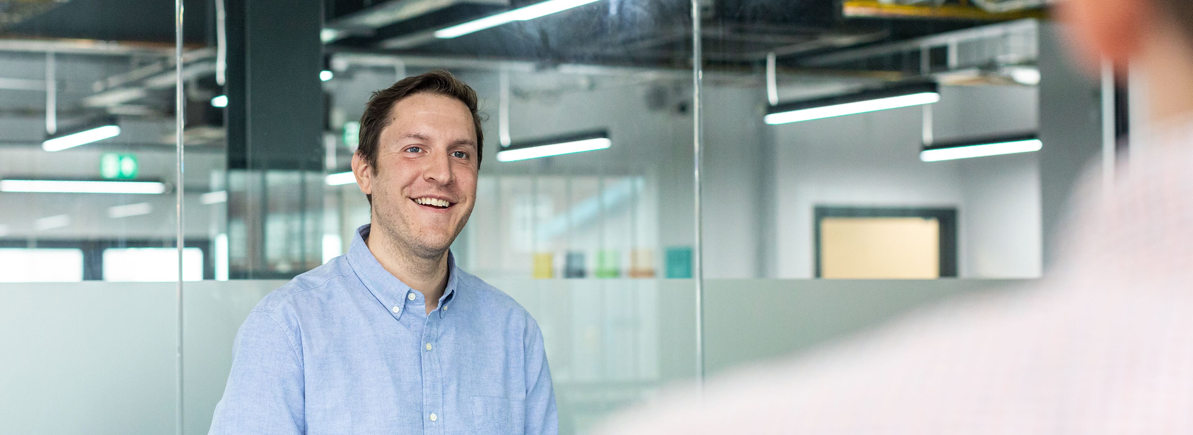 A man in a light blue shirt is smiling in an office setting, standing near a glass partition with fluorescent lights in the background. Another person's shoulder is visible in the foreground.