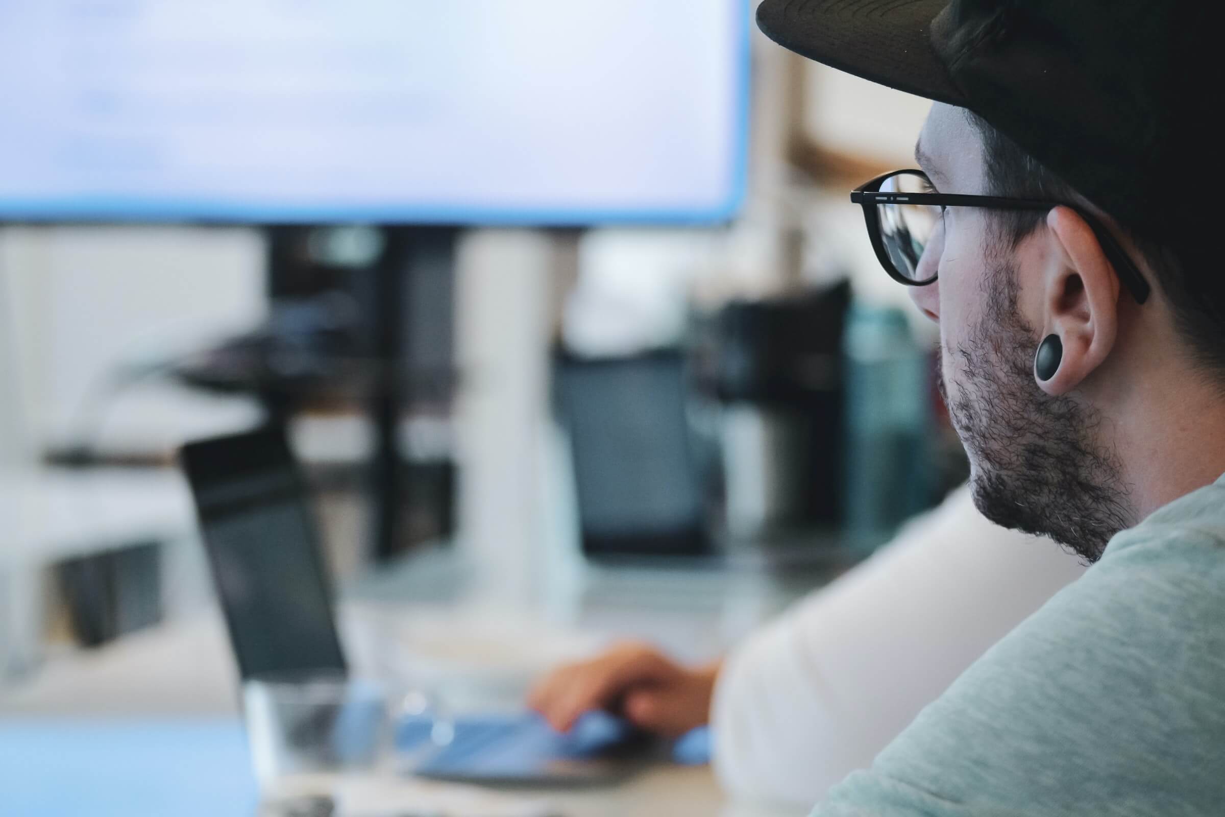 A person wearing glasses and a cap is using a laptop, focusing on the screen. An out-of-focus monitor is visible in the background.