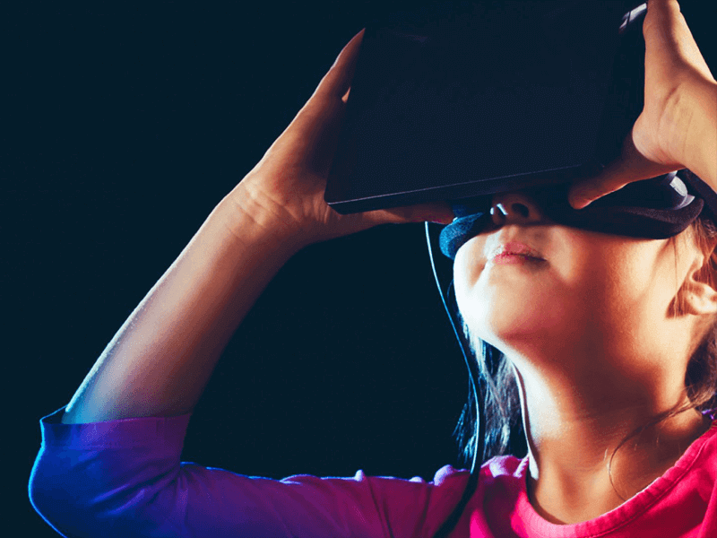 A young person with long hair, wearing a pink shirt, holds a VR headset against their eyes, immersed in a virtual experience against a dark background.