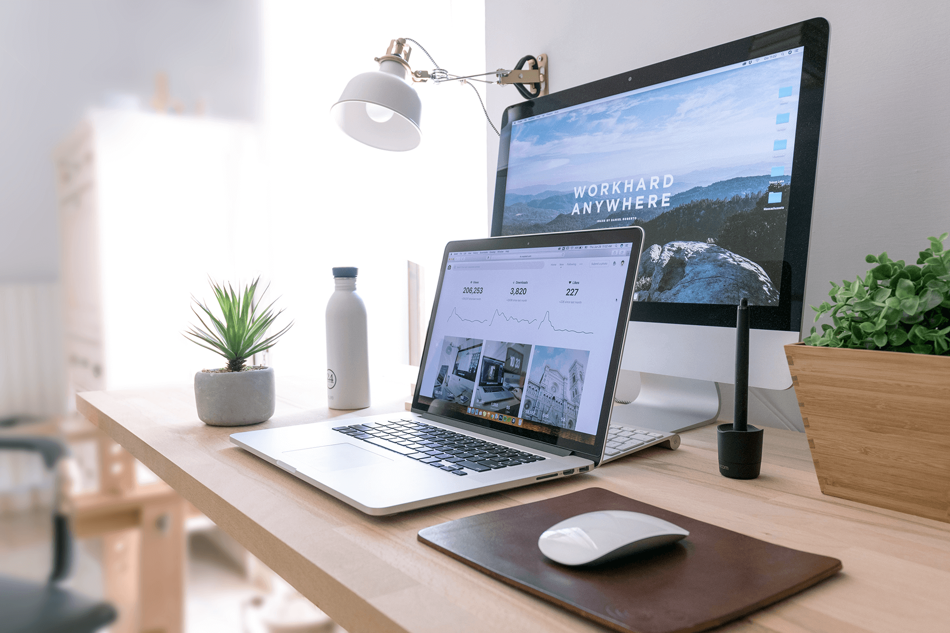 A desk with a laptop, desktop monitor displaying "WORK HARD ANYWHERE", potted plants, water bottle, tablet pen, and desk lamp.