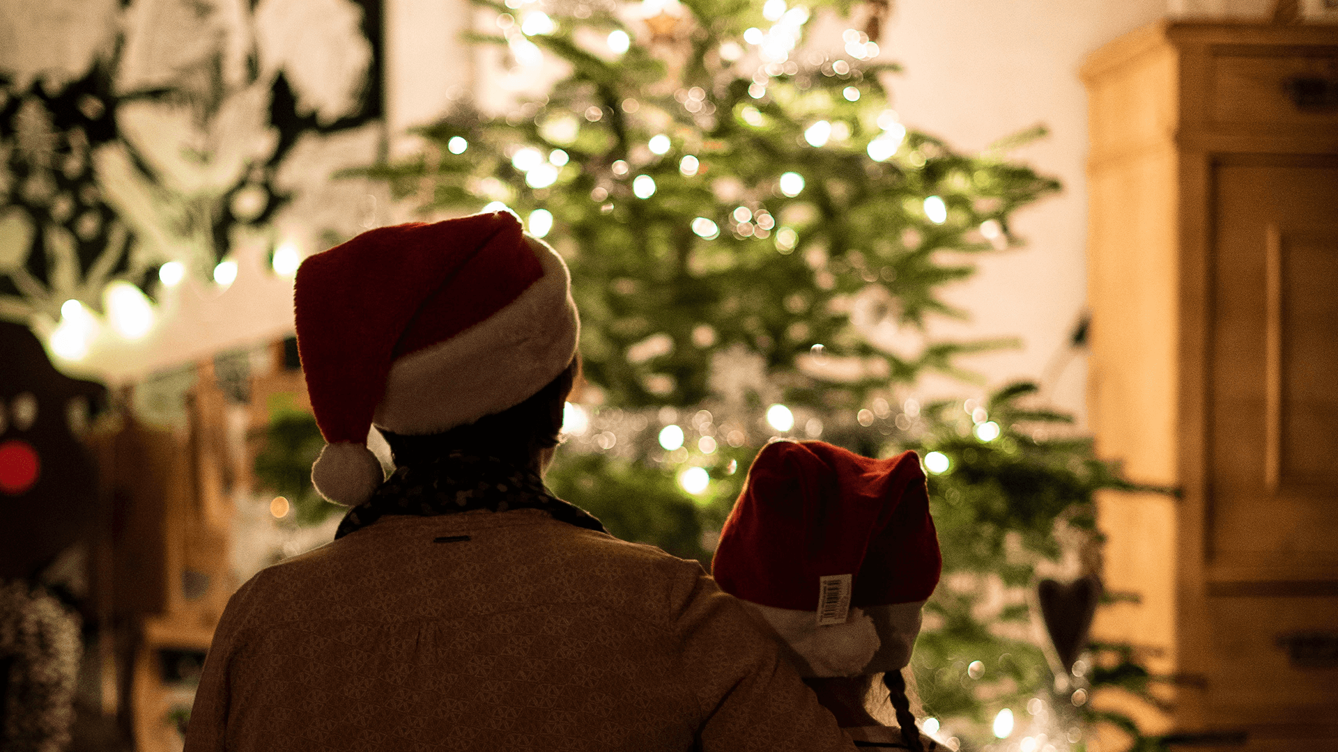 Two people wearing Santa hats stand closely together, facing a decorated and lit Christmas tree.