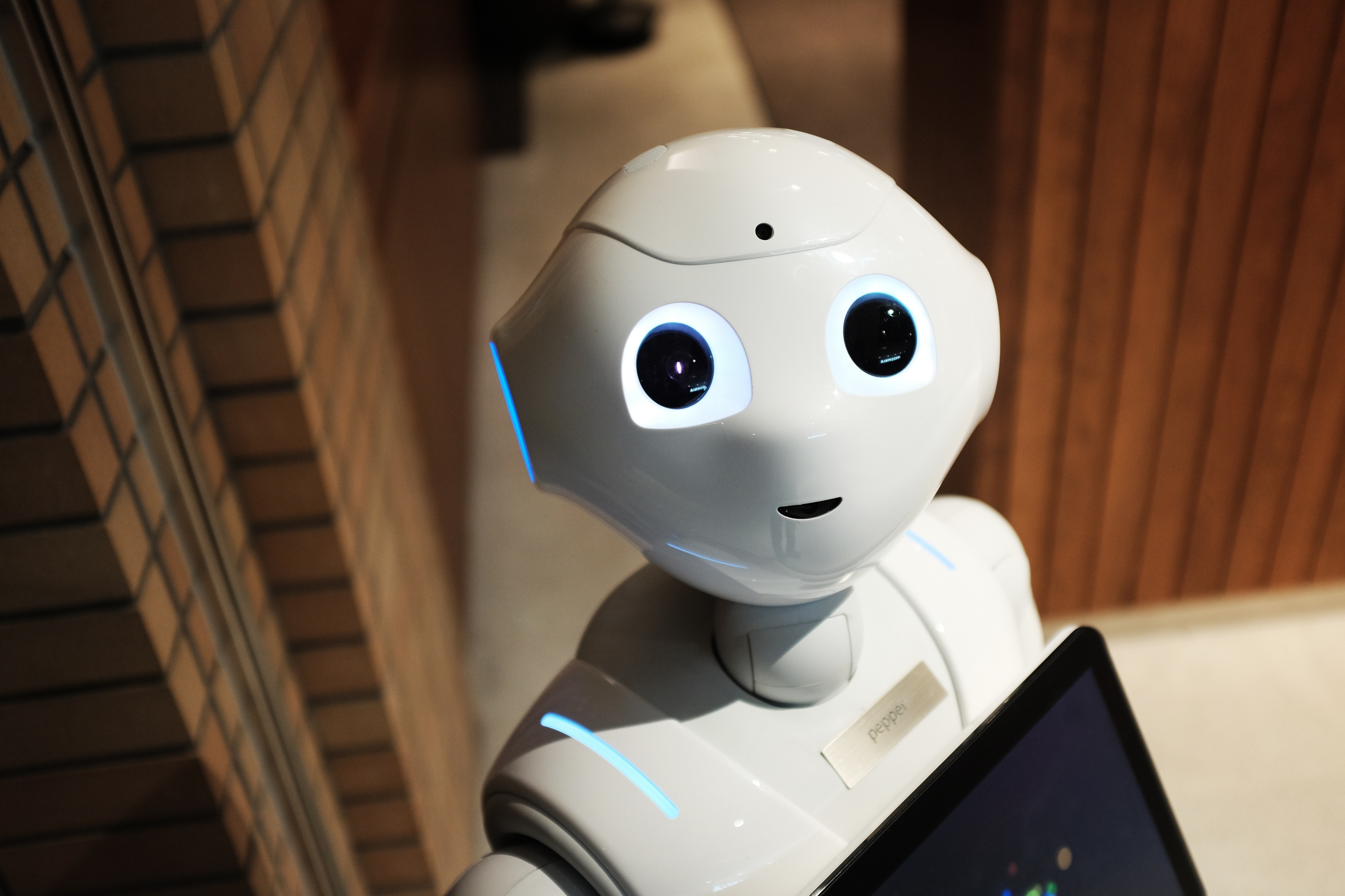 A humanoid robot with white plastic casing and large, black eyes is holding a tablet, standing indoors near a brick wall.