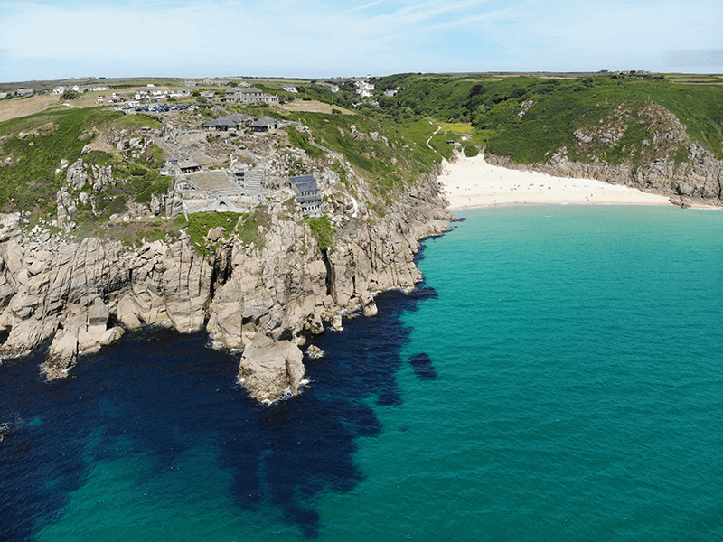 Aerial view of a coastal landscape with rocky cliffs, a sandy beach, and turquoise ocean water. There are scattered buildings and greenery on top of the cliffs.