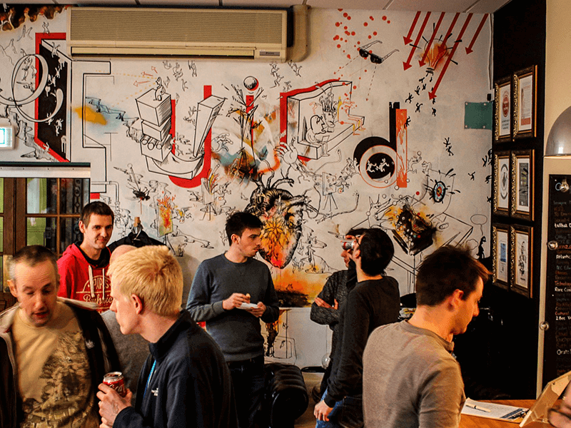 Several people are socializing in a room with a colorful, graffiti-style mural covering one wall. There are framed pictures hanging on the adjacent wall.