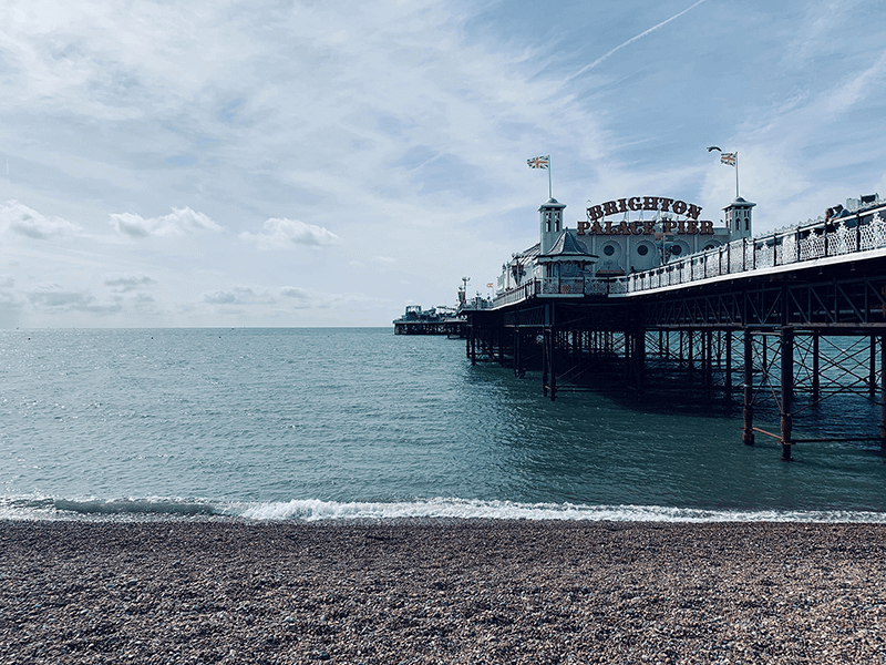 A view of Brighton Palace Pier extending over the sea, with a pebble beach in the foreground and a cloudy sky in the background.