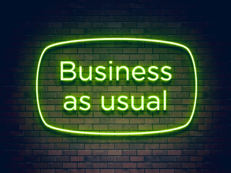 Neon green sign reading "Business as usual" against a dark brick wall background.