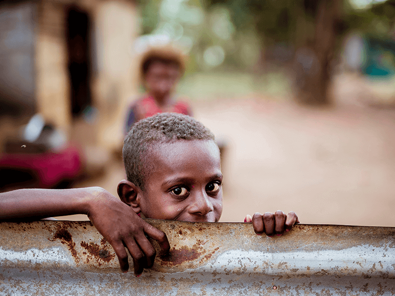 A child peeks over a rusted metal sheet, holding onto the top edge, with a blurred background of another person and a rural setting.