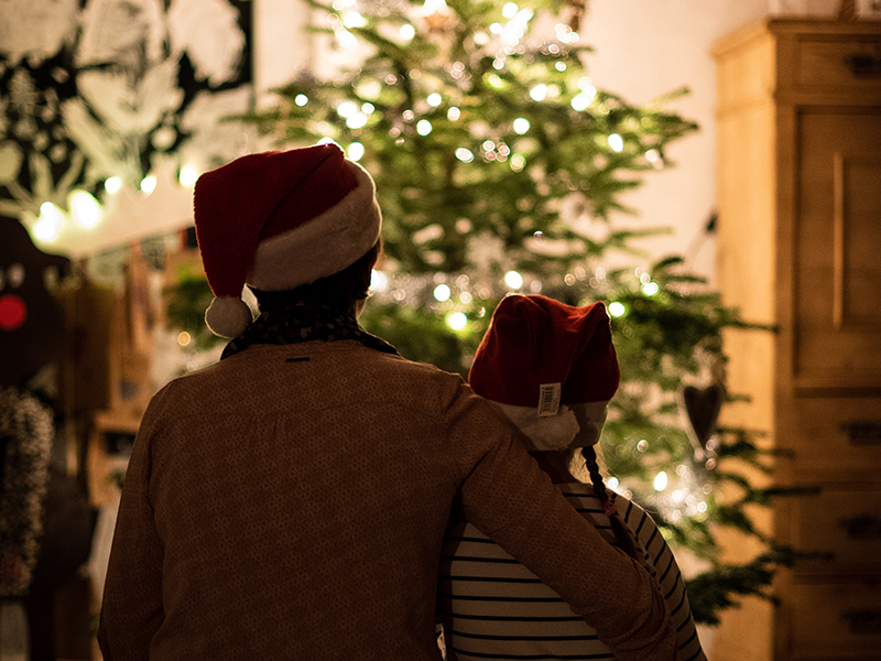 Two people wearing Santa hats stand close together, facing a lit Christmas tree in a cozy, dimly lit room.