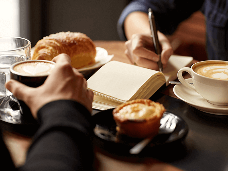 Two people engage in offline networking over coffee and pastries while one writes in a notebook. Both have cups of coffee with latte art, surrounded by croissants and other delectable pastries, highlighting why face-to-face communication matters in a digital era.