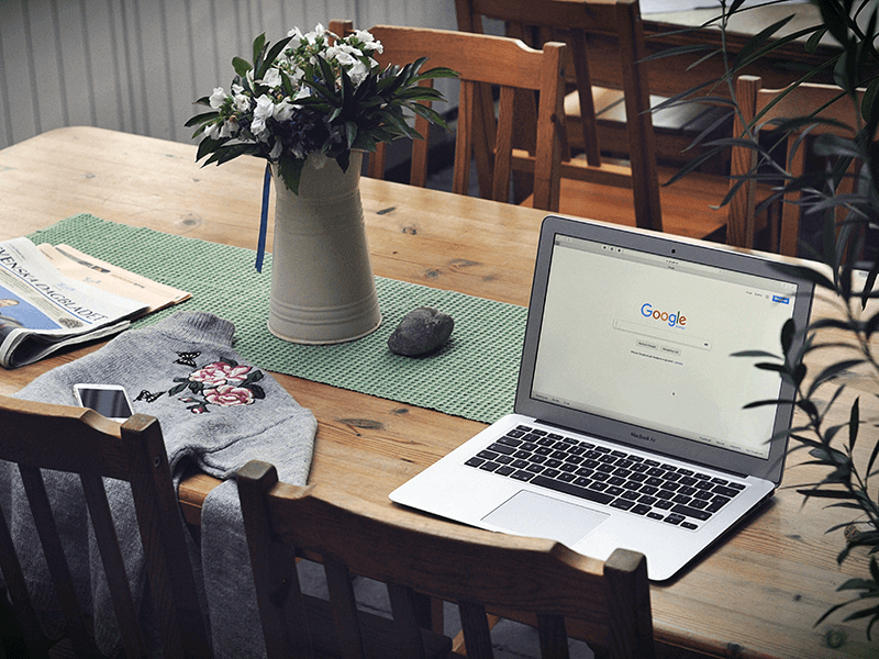 A wooden table with a laptop displaying Google's homepage, a smartphone, a newspaper, a gray sweatshirt with flowers, a green table runner, and a vase with white flowers.