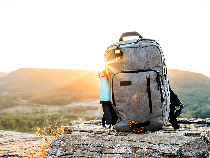 A gray backpack and a blue water bottle are placed on a rock ledge at sunrise, overlooking a valley and distant hills.