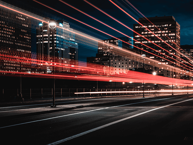 A nighttime cityscape with light trails from moving vehicles, tall buildings with lit windows in the background, and a clear, dark sky.