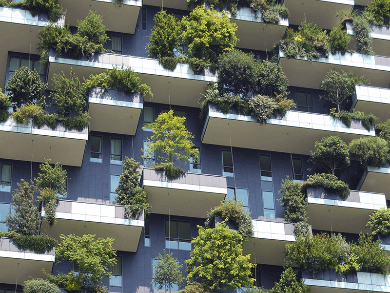 A modern multi-story building with balconies adorned with lush green plants and trees, creating a vertical garden effect.