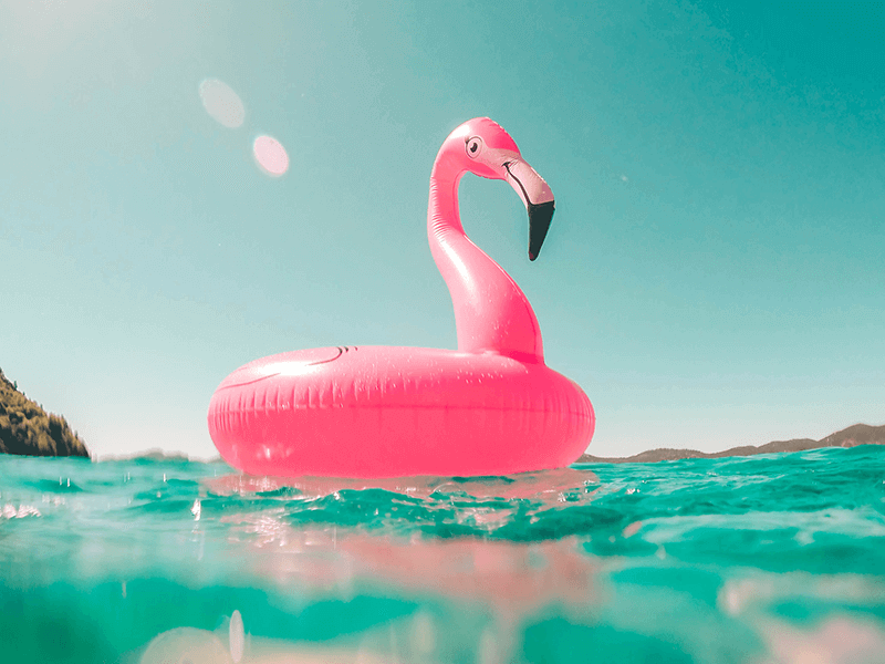 A pink flamingo-shaped inflatable float sits on clear turquoise water under a bright blue sky.