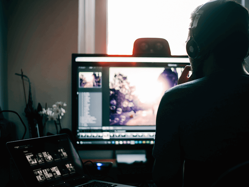 A person wearing headphones works on a desktop computer and a laptop, editing a photo with sunlight shining through a window in the background.