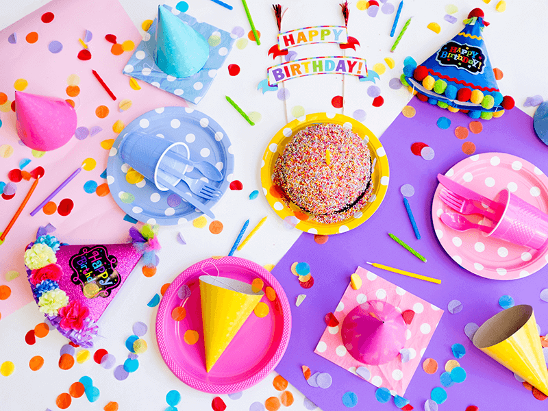 A colorful birthday party setup with decorated hats, scattered confetti, polka dot plates and napkins, plastic utensils, and a sprinkled cake. The scene is vibrant and festive.