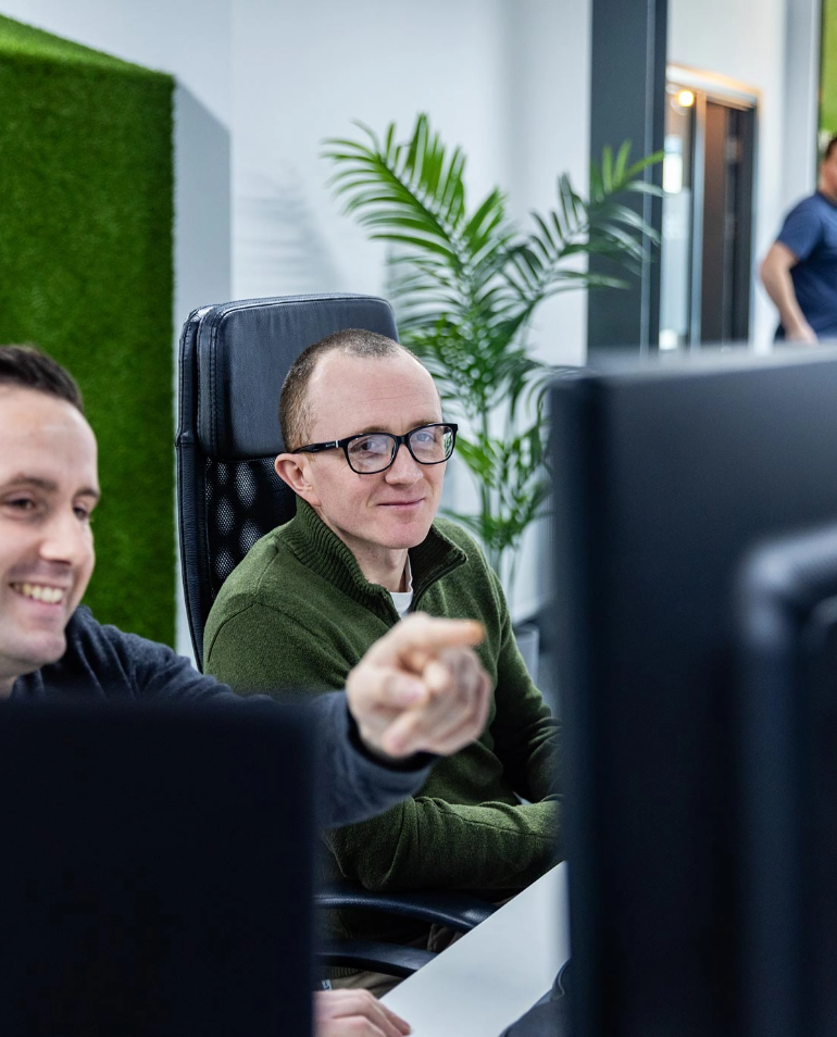 Two people are sitting at desks with computer monitors. One person, smiling, points at something on their screen while the other person, wearing glasses, looks on attentively. An out-of-focus person is in the background.