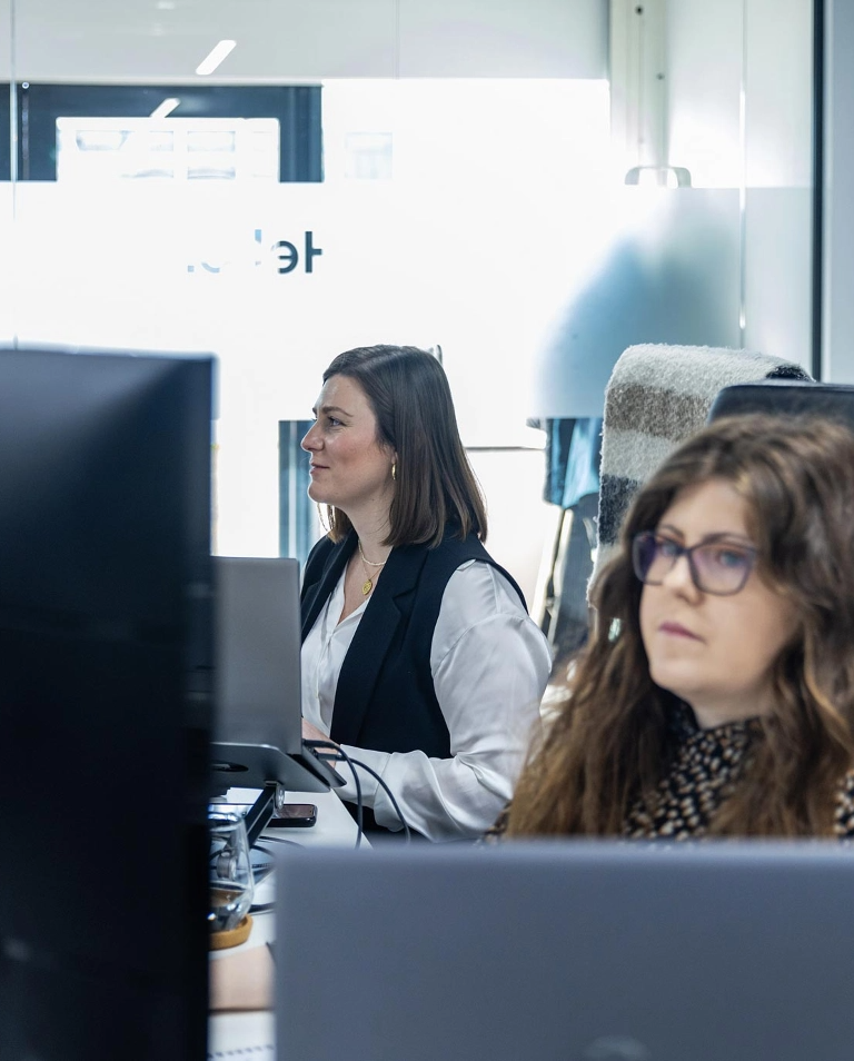 Two women are working at their desks in an office, focused on their computer screens. The background includes frosted glass and a window.