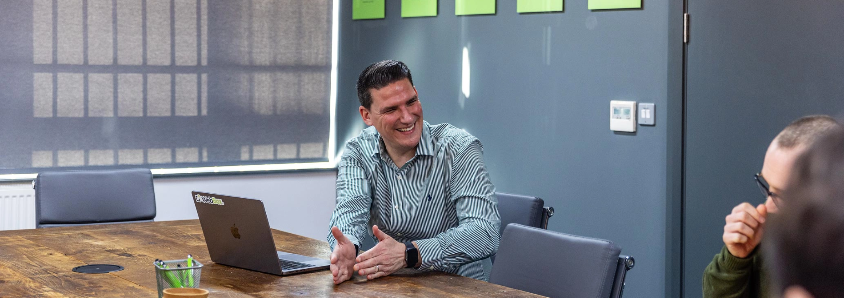 A man in a light green shirt is smiling and gesturing while sitting at a conference table with a laptop. Another person is partially visible in the foreground. The room has a grey wall with notes on it.