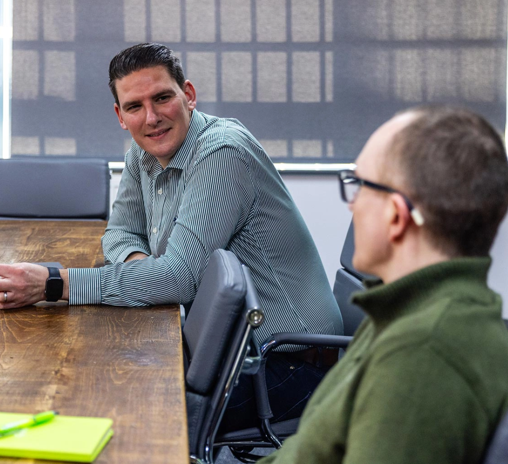 Two individuals are seated at a wooden conference table, engaged in conversation. One is wearing a striped shirt, while the other wears a green shirt and glasses. A yellow notebook is on the table.