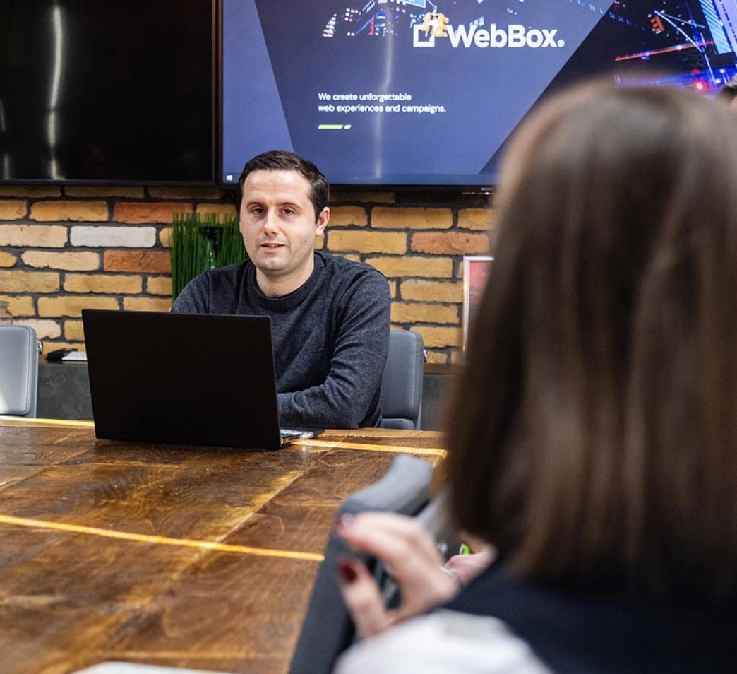 A man sits at a wooden table with a laptop in front of him, facing a woman. A screen behind him displays the word "WebBox" and other text.