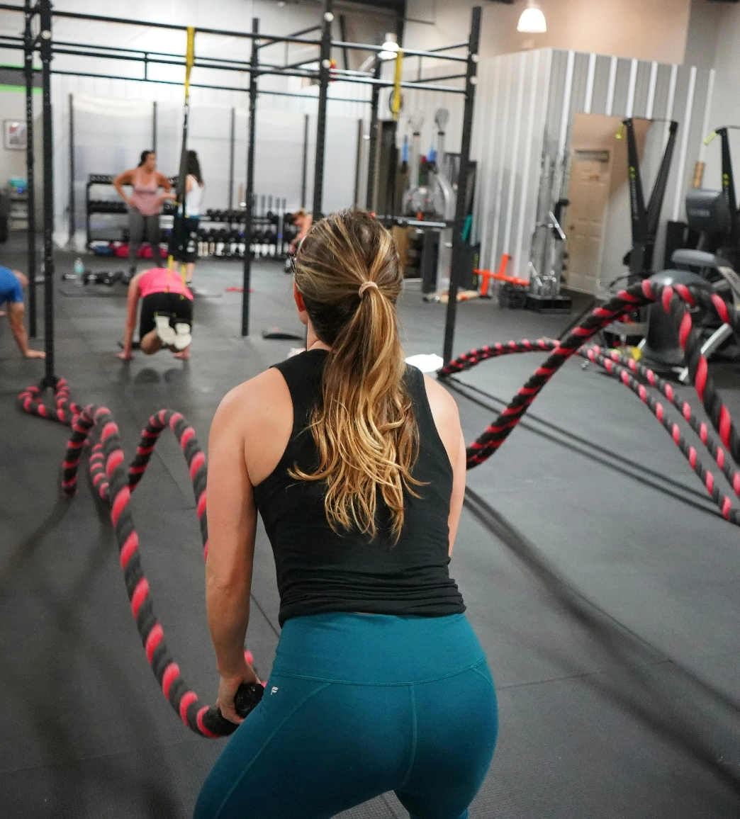 A woman exercises with battle ropes in a gym, while other individuals perform various workouts in the background.