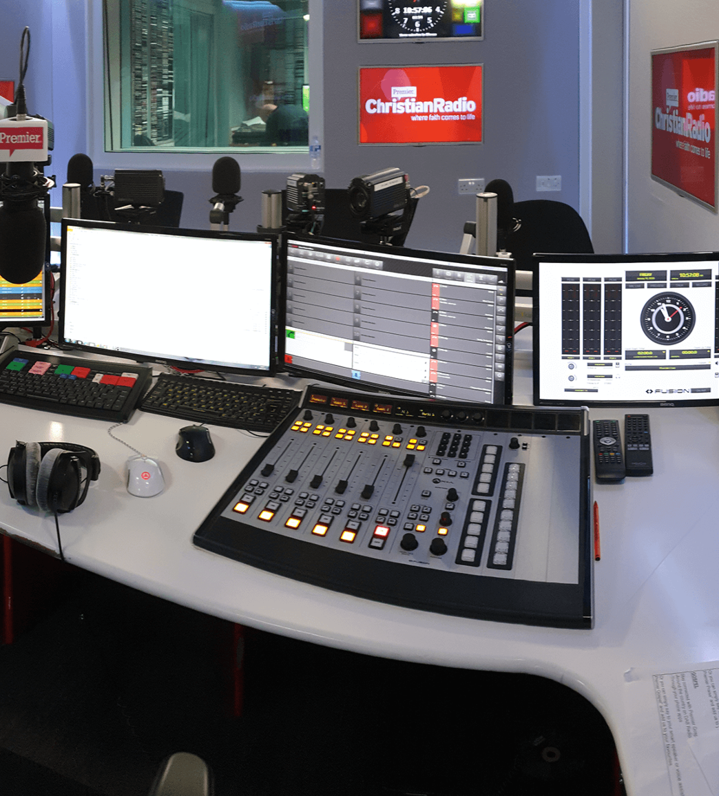 A radio station studio setup with a mixing console, multiple computer monitors, a keyboard, headphones, and microphones. Walls feature Christian Radio logos.