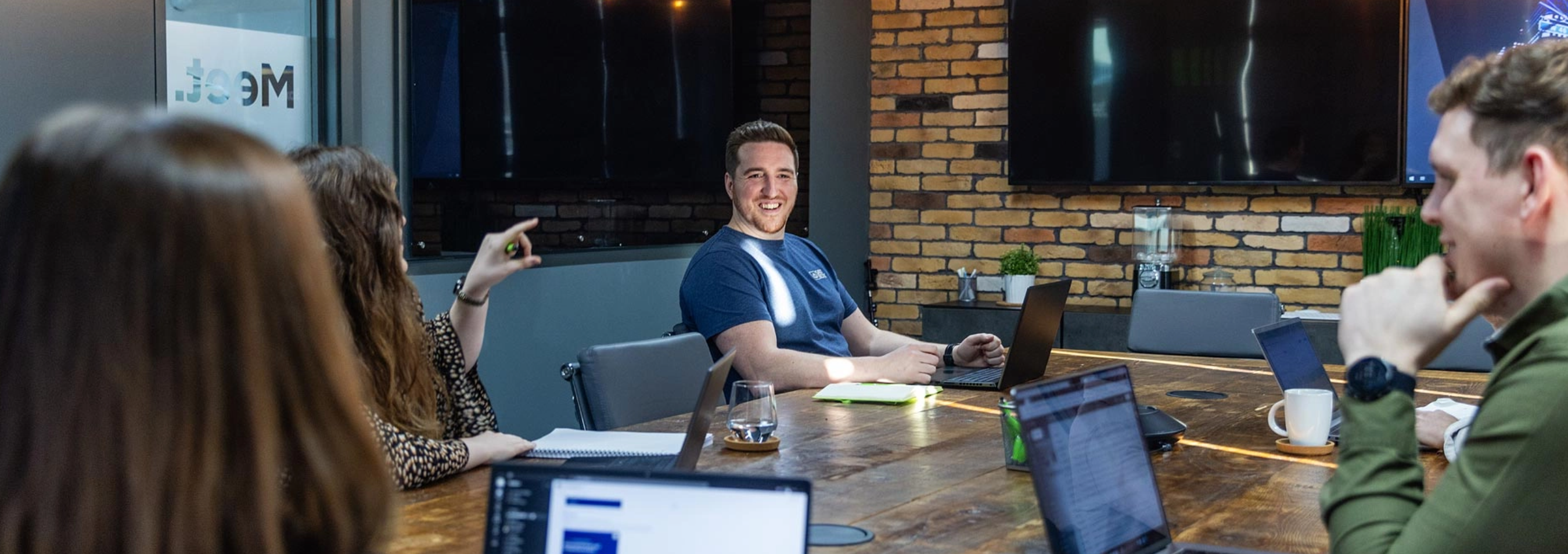 A group of people sit around a large wooden conference table with laptops, having a discussion. One person smiles while others are engaged. Brick wall and large screens are in the background.