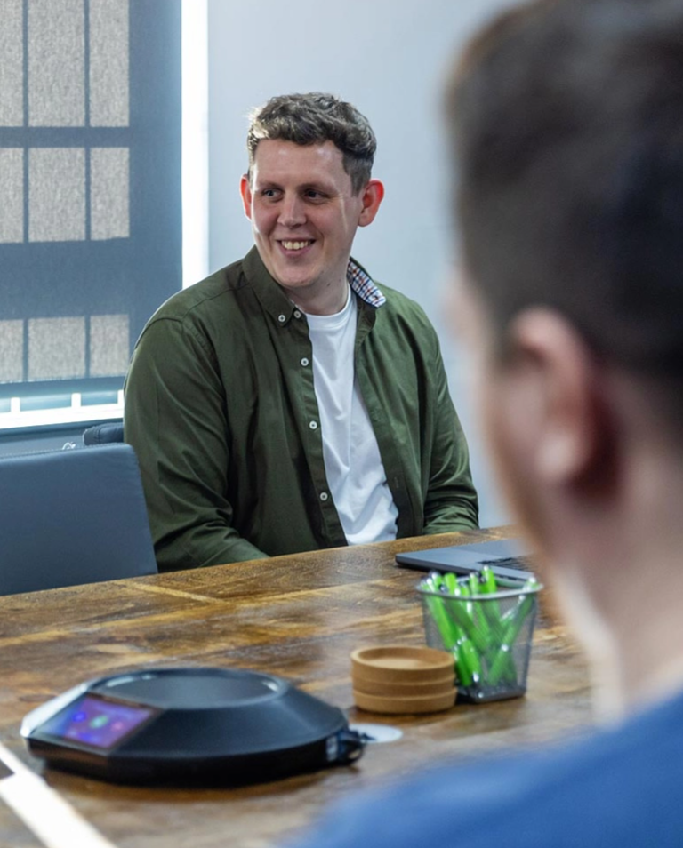 A man in a green shirt sits at a wooden table with office supplies and a conference speaker, smiling and engaging in conversation during a meeting.