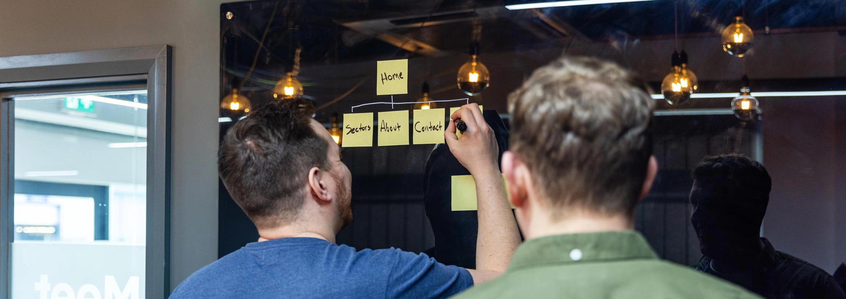 Two individuals are seen from behind, collaborating on a flowchart with sticky notes on a glass board, in a modern office space with hanging lights.