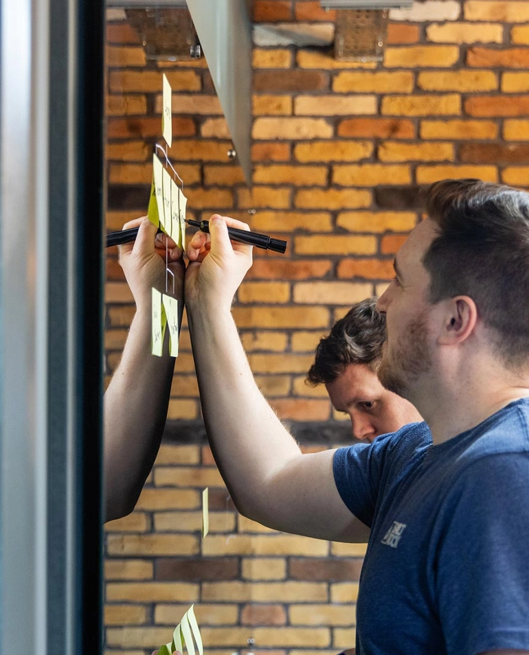 Two adults in a room with a brick wall, writing on large sticky notes attached to a glass surface. One person is focused on writing, while the other looks at the notes.