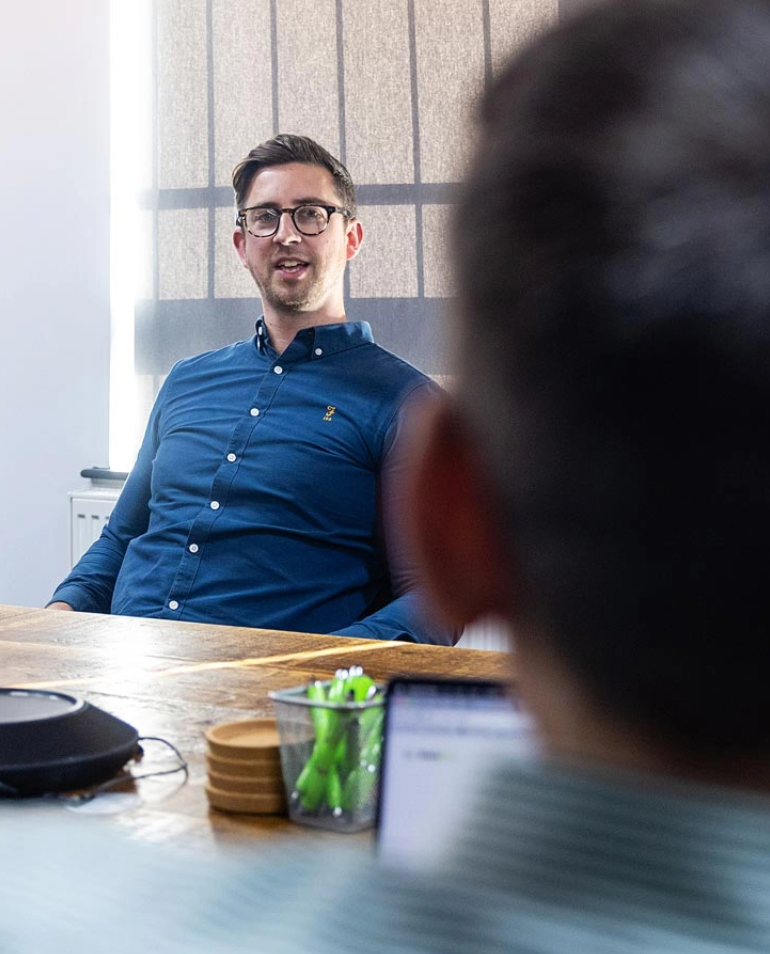 A man in a blue shirt is seen talking during a meeting, seated at a wooden table, with another person partially obstructing the foreground.