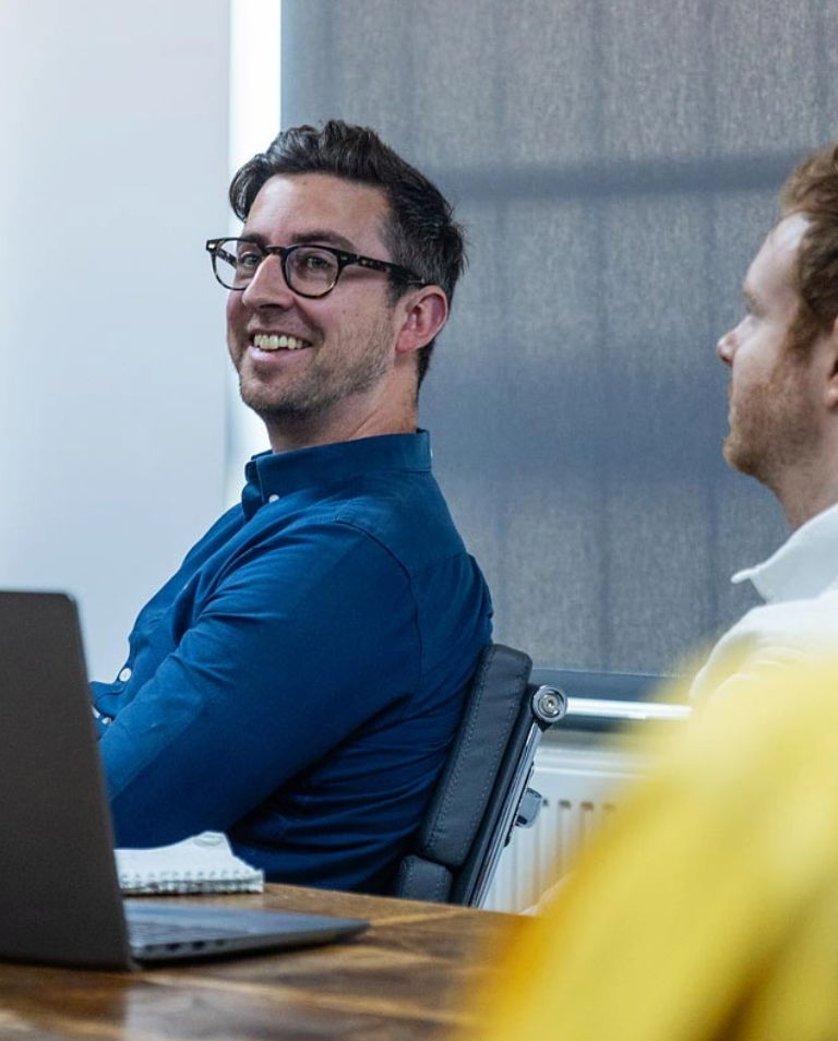 Two men are sitting at a table in an office environment. The man on the left is smiling and wearing glasses and a blue shirt. A laptop and notepad are in front of him.